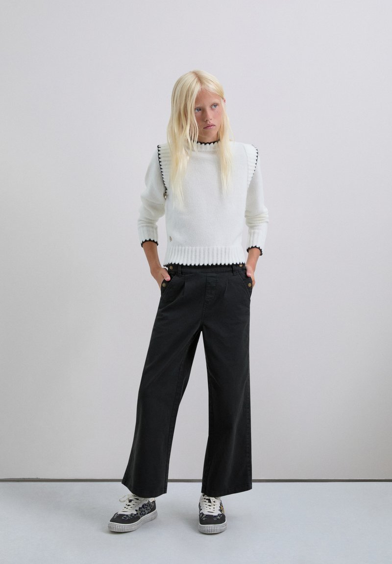 Suéter blanco de punto con terminaciones en negro en forma de concha, combinado con pantalones negros de corte holgado y zapatillas con estampado. Fondo minimalista.