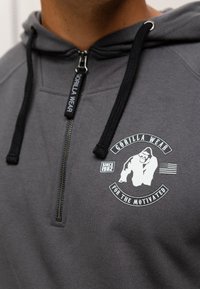 Grauer Kapuzenpullover mit Reißverschluss, schwarzen Kordelzügen und einem grafischen Logo. Mit dem Text "Gorilla Wear" und einem Design mit einem Gorilla und der amerikanischen Flagge.