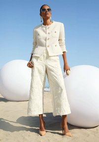 Witte textuur blazer met gouden knopen, gecombineerd met brede crèmekleurige broek met bloemen borduurwerk. Vergezeld van nude hoge hakken.