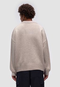 Beżowy oversize sweter z prążkowanym dołem, opadającymi ramionami i miękką fakturą. Noszony z ciemnymi spodniami cargo, widok z tyłu.