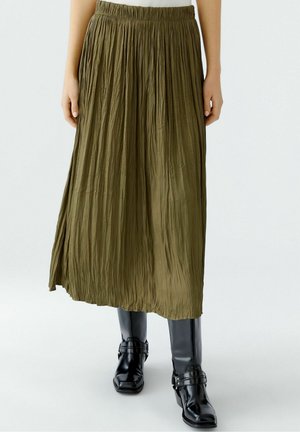 Olivengrøn krøllet midi-nederdel kombineret med sorte læderstøvler til midt på læggen på en person, der står mod en ensfarvet baggrund.