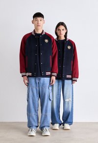 Marineblå og bordeaux varsity jakker med hvide stribede kraver, udstyret med guld logo patches; bæres med løse denim bukser og sneakers.
