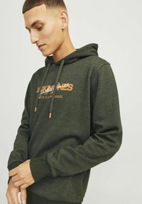 Sudadera de color verde oscuro en una tela suave con puños de canalé. Cuenta con cordones con detalles en marrón y un logotipo de la marca impreso en blanco en la parte frontal.