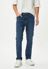 Jeans de mezclilla azul con un diseño ajustado, cinco bolsillos y detalles de costura visibles, combinados con zapatillas blancas sobre un fondo simple.