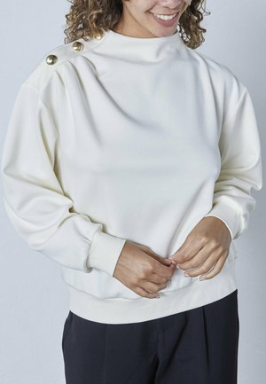 Hvid sweatshirt med høj hals og guldknapdetaljer på skulderen. Blødt stof, afslappet pasform og ribbede manchetter og kant.