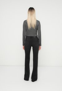 Kobieta z długimi blond włosami stoi przodem do białej ściany, ubrana w szary top z długim rękawem i czarne dżinsy w stylu bootcut.