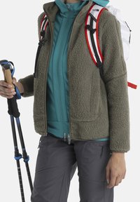 Giacca in pile verde oliva con una finitura texturizzata, indossata sopra una maglietta blu con zip. I pantaloni grigi e i bastoni da trekking completano il look.