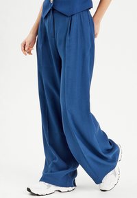 Pantalon à jambes larges en tissu bleu foncé, avec des plis et une coupe décontractée. Associé à des baskets blanches, mettant en avant le confort et le style.