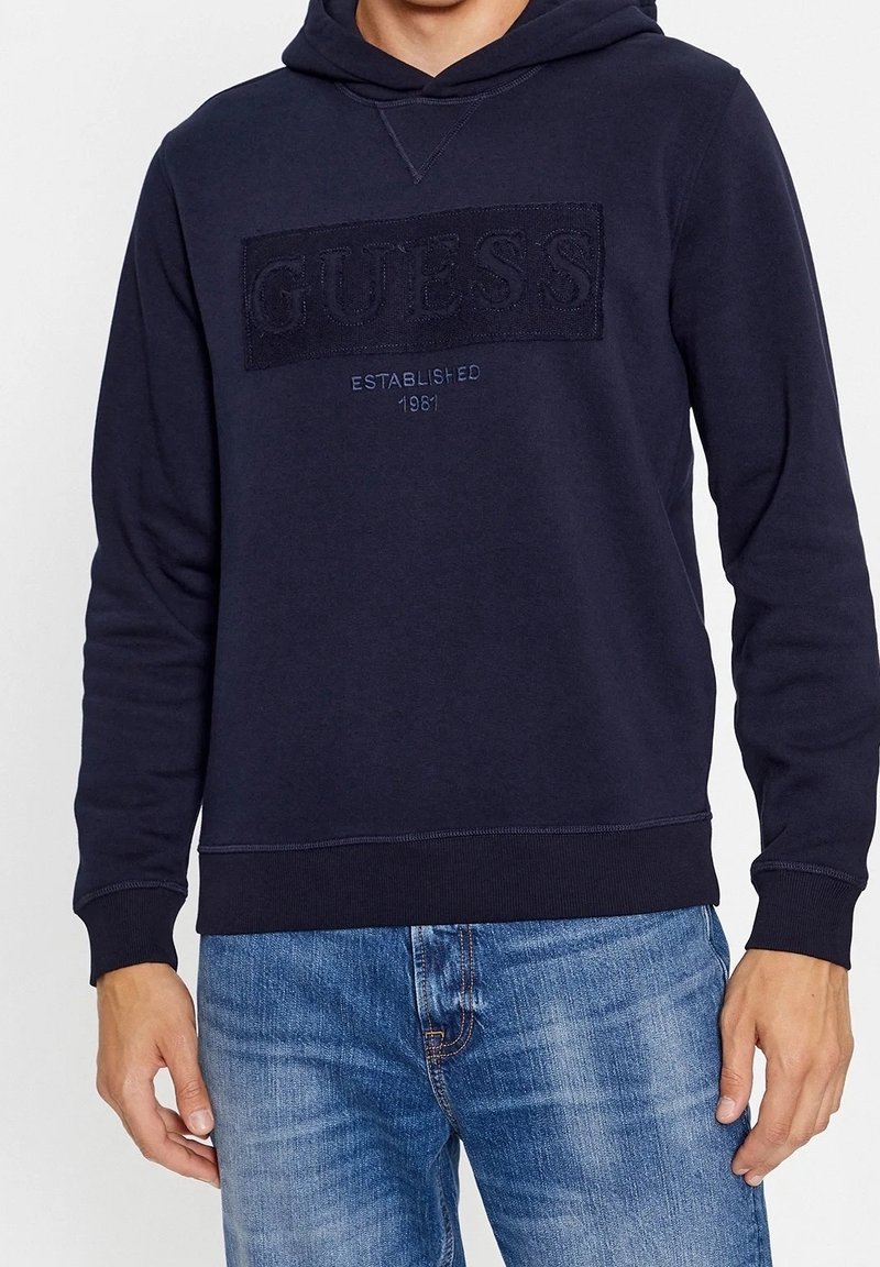 Sudadera en azul marino hecha de tela suave, con un logotipo texturizado de "GUESS" en el pecho, y puños y dobladillo acanalados para mayor comodidad.