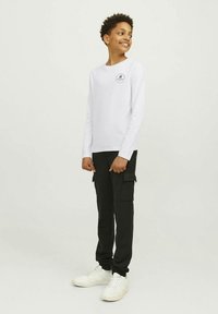 Valge pikkade varrukatega T-särk ümmarguse logoga, millel on mustad küljetaskutega cargo-püksid ning valged tossud.