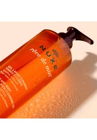 Orange flaska med Nuxe duschgel som har en pump-topp, med en texturerad yta och kondensdroppar, märkt för användning på ansikte och kropp.