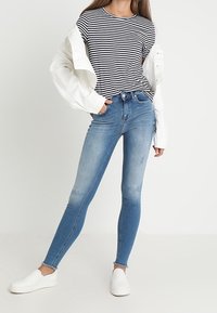 Úzké modré džíny s vyblednutím na stehnech, zkombinované s pruhovanou černou a bílou košilí a bílou oversized bundou. Bílé slip-on boty.