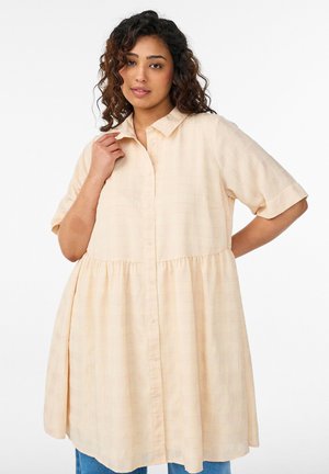 Woman with curly hair wearing a loose, pale peach button-up dress with short sleeves and a gathered waist, posing against a white background.