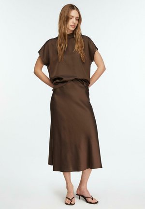 Kobieta z długimi włosami, ubrana w brązową satynową bluzkę z krótkim rękawem i pasującą spódnicę midi, stojąca z rękami za plecami w czarnych sandałach na obcasie.