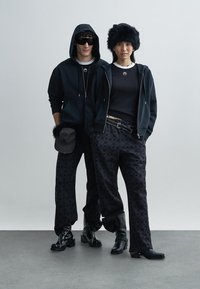 Melns krekls ar kapuci un drukātas bikses, abiem ir faktūras raksts. Modelis valkā kažokādas cepuri un saulesbrilles. Apavi ir melni un spīdīgi.