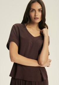 Krótka brązowa bluzka typu v-neck z krótkimi, podwiniętymi rękawami i luźnym krojem. Wykonana z gładkiego materiału, ma subtelny wzór i opada tuż poniżej talii.