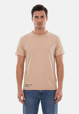 Uomo con capelli corti e scuri che indossa una semplice t-shirt beige a maniche corte e jeans blu, in piedi contro uno sfondo chiaro.