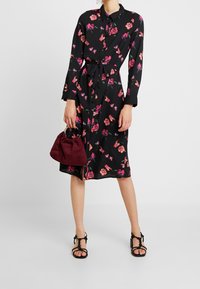 Robe florale noire à manches longues et ceinture à nouer, arborant des motifs floraux roses et verts. Associée à un sac à main bordeaux et des sandales noires.