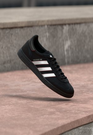 Baskets Adidas noires avec des bandes blanches et un marquage "Messi" flottant au-dessus d'une surface en pierre et d'un mur gris en arrière-plan.