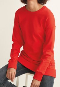 Personne portant un pull à manches longues rouge vif et un jean foncé, assise sur un tabouret blanc avec des cheveux bouclés visibles.