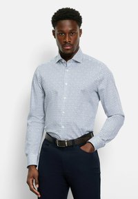 Weißes Langarmhemd mit blauem, kreisförmigem Muster, Button-Down-Kragen und schmaler Passform, kombiniert mit marineblauen Hosen und einem braunen Gürtel.