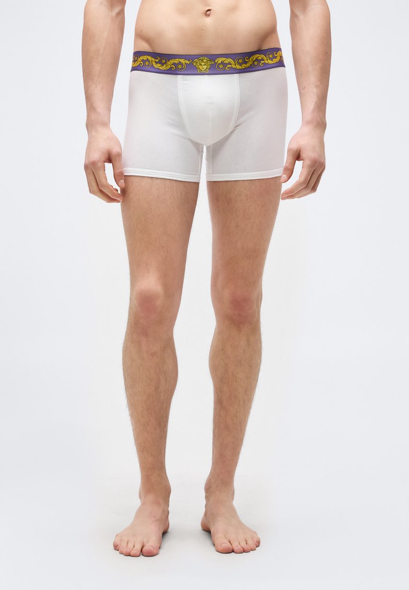 Άνδρας μοντέλο που φοράει λευκά ανδρικά εσώρουχ α μποξεράκια με ζώνη μέσης σε μοτίβο μωβ και χρυσό, με λογότυπο κεφαλής Μέδουσας, στέκεται ξυπόλητος σε απλό λευκό φόντο.