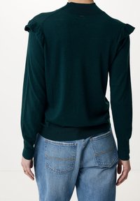 Person som bär en mörkgrön långärmad tröja med volanger på axlarna och ljusblå denimjeans, sedd bakifrån.