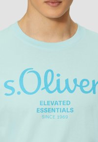Hele sinine puuvillane T-särk, millel on kaubamärk "s.Oliver" julgete türkiissiniste tähtedega ja all "ELEVATED ESSENTIALS" tekst. Ümar kaelus.