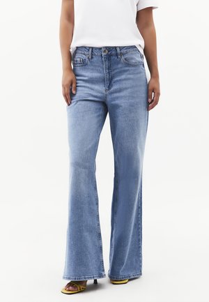 Person iført lyseblå jeans med høj talje og bredde forneden, hvid T-shirt og gule åbne tå-sandaletter med hæl, stående mod hvid baggrund.