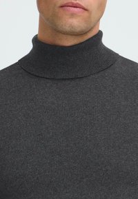 Uomo che indossa un maglione a collo alto a coste grigio scuro, primo piano sul collo e sulla parte bassa del viso su uno sfondo neutro.