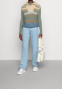Dzianinowa kamizelka w wielokolorowe paski w odcieniach zieleni i niebieskiego, zestawiona z jasnoniebieskimi jeansami o prostych nogawkach i białymi trampkami, trzymająca kremową torebkę.