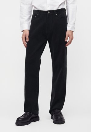 Pantalon droit en velours côtelé noir porté avec des chaussures en cuir noires et une chemise blanche à manches longues boutonnée.