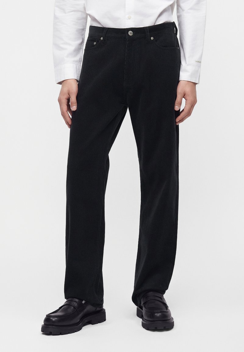 Pantalon droit en velours côtelé noir porté avec des chaussures en cuir noires et une chemise blanche à manches longues boutonnée.