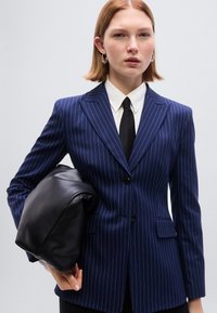 Μπλε ναυτικό σακάκι με ρίγες, καλοκομμένη εφαρμογή, πέτο με σχισμή και δύο εμπρόσθιες τσέπες. Κρατιέται μία μαύρη δερμάτινη τσάντα κάτω από το ένα χέρι.