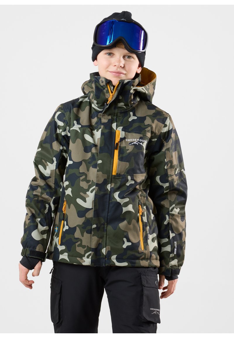 Kamouflage skidjacka i grön, svart och vit med orange dragkedjor. Har en huva, fickor och logodetaljer på bröstet och ärmen.