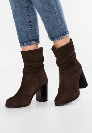 Braune Wildleder-Stiefeletten mit Blockabsätzen, getragen zu blauen, kurz geschnittenen Jeans auf weißem Boden.