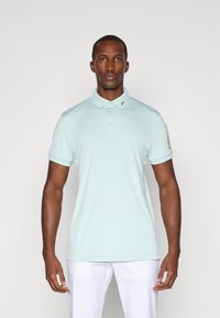 Camisola polo azul-claro com colarinho, três botões e um logotipo discreto na gola. Mangas curtas com uma textura suave.
