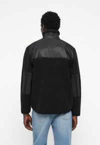 Black fleece jacket with glossy black nylon shoulder and sleeve accents. Features a high collar and relaxed fit. Back view shows texture contrast.