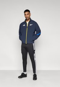 Ναυτικό αθλητικό μπουφάν με κίτρινο φερμουάρ και μπλε ρίγες, με λογότυπο. Συνδυάζεται με μαύρο στενό παντελόνι με λευκές πλαϊνές ρίγες και μαύρα παπούτσια.