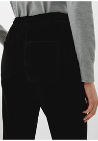 Pantalon en velours côtelé noir avec une surface texturée, ceinture élastique et deux poches arrière plaquées. Associé à un haut à manches longues gris clair.