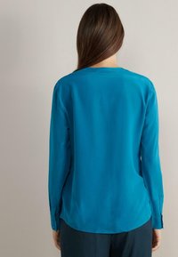 Turquoise long-sleeve blouse with a smooth texture, round neckline, and buttoned cuffs. Tailored fit with a curved hem at the back.