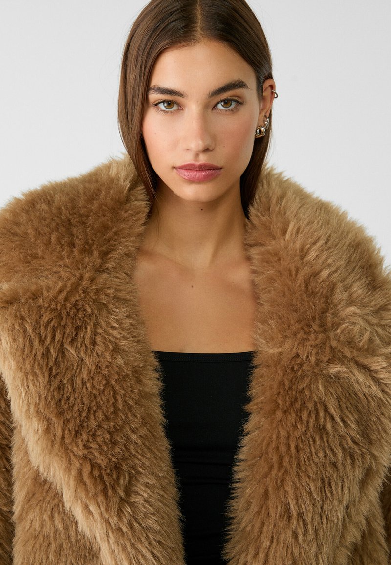 Cappotto in pelliccia sintetica marrone con un colletto voluminoso. La texture è morbida e soffice, abbinata a un top nero aderente sottostante.