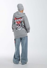 Felpa grigia con una grafica di teschio e cuore sul retro, abbinata a jeans larghi blu chiaro con una patch floreale colorata.