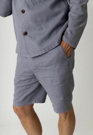 Mężczyzna ubrany w jasnoszarą koszulę na guziki i pasujące spodenki, z jedną ręką w kieszeni, na jednolitym tle.