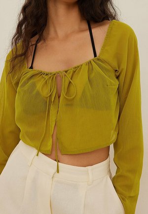 Kobieta ubrana w przezroczystą, oliwkowozieloną krótka bluzkę z długimi rękawami, związaną na biuście, nałożoną na czarny stanik, w połączeniu z białymi, wysokim stanem spodniami.