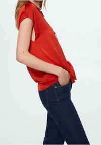 Blouse rouge à manches courtes avec une coupe ample, présentant un design ouvert sur les côtés, associée à un jean bleu foncé. Texture de tissu lisse.