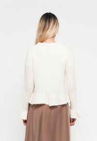 Frau mit langen blonden Haaren trägt einen weißen strukturierten Pullover mit Rüschensaum und -ärmel, kombiniert mit einem braunen Rock, von hinten zu sehen.