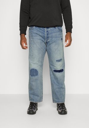 Mand iført lyseblå jeans med mørkeblå lapper på knæene, sorte sneakers og en sort langærmet skjorte står mod en hvid baggrund.