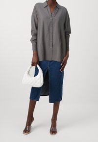 Chemise grise à boutons avec une coupe décontractée, jupe en denim bleu avec une fente à l'avant, sandales à talons transparents, et sac à main blanc matelassé.