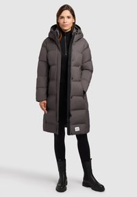 Padded gray coat with a hood, closed with buttons, featuring a logo patch at the hem, paired with black pants and knee-high boots.
